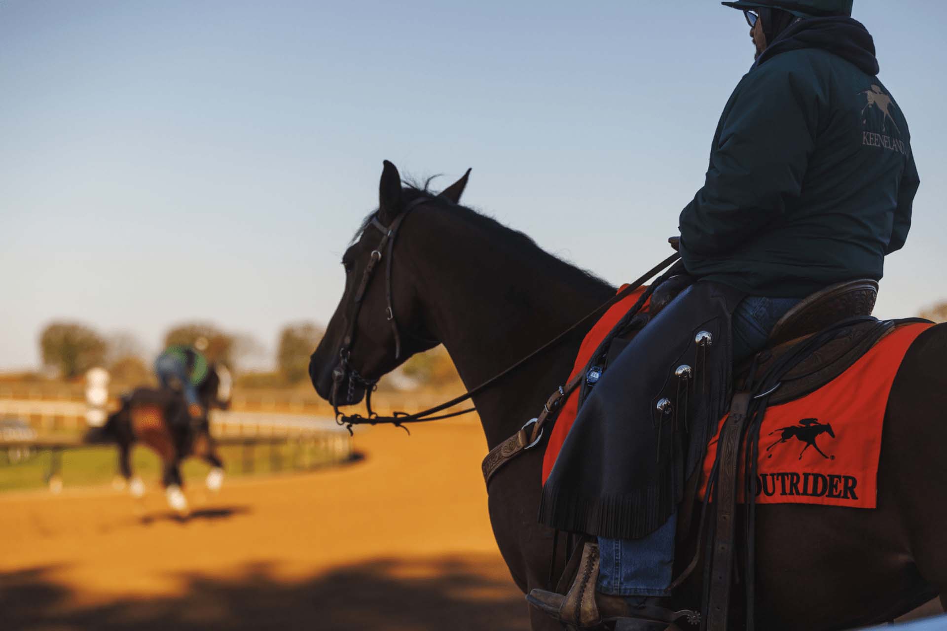 A candid photo from the side of a man riding a brown horse. There is an orange flag on the horse that reads “Outrider”.